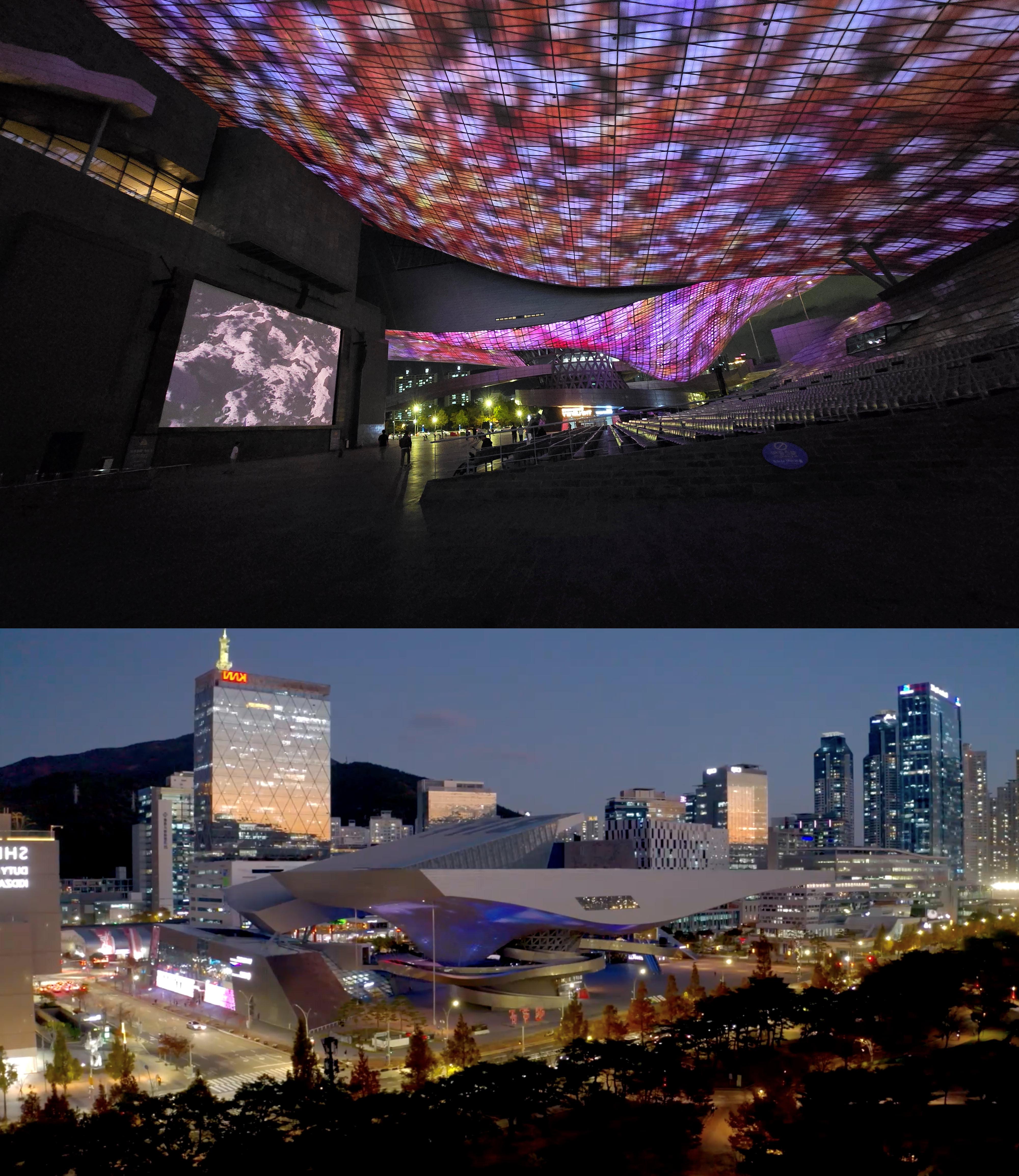  A futuristic architectural space with a large, curved LED ceiling displaying vibrant, abstract visuals in red, purple, and blue hues. The darkened interior has a few people walking, and a large screen on the wall shows a black-and-white image of Earth's surface. Below, a second image shows a cityscape at dusk with a modern, spaceship-like building illuminated in soft blue light, surrounded by high-rise buildings and streetlights. The urban environment is lively, with cars and people visible in the streets.